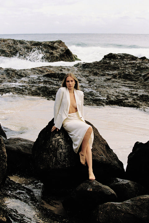 Model wearing white linen blazer and cream silk slip skirt posing on a rock by the beach