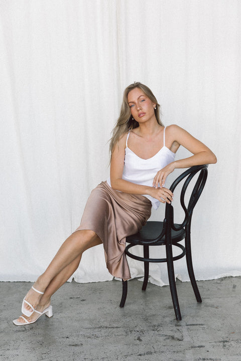 Model wearing a white silk camisole and taupe silk slip skirt posing on a chair in a studio