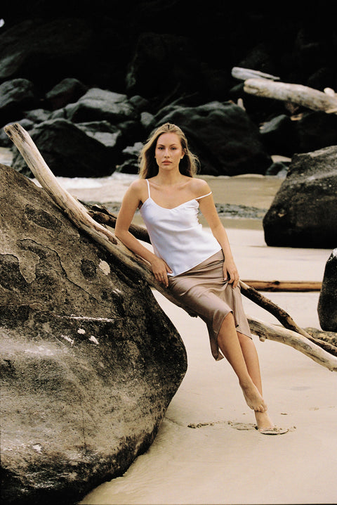 Model wearing white silk camisole and taupe silk slip skirt posing on a rock by the beach